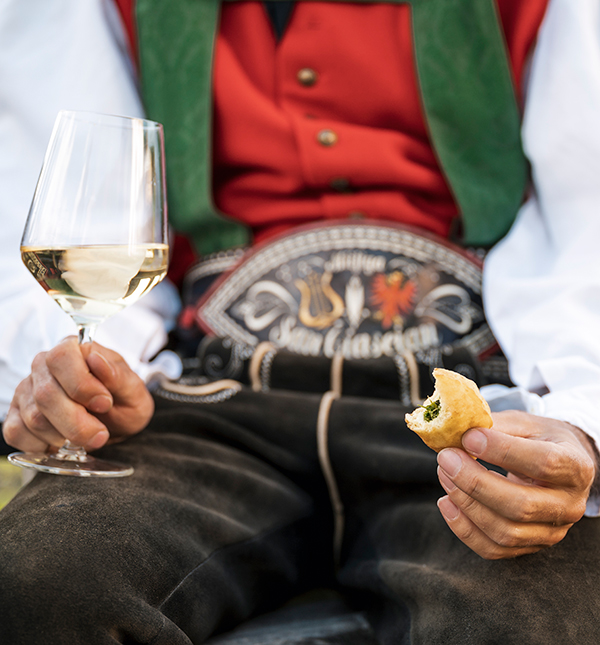 ein Mann in Tracht während einer traditionellen Veranstaltung mit einem Weinglas und einem Krapfen in der Hand>